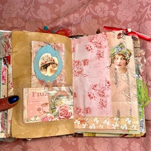May include: Open journal with vintage-style pages. Features include a blue framed portrait of a woman in a hat, a floral print, and a pink and white floral design. The journal is open on a pink floral patterned background.