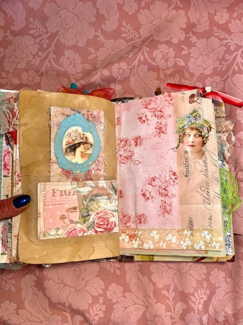 May include: Open journal with vintage-style pages. Features include a blue framed portrait of a woman in a hat, a floral print, and a pink and white floral design. The journal is open on a pink floral patterned background.