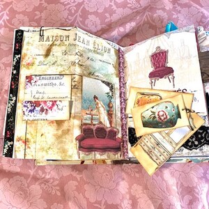 May include: Open journal with vintage-style pages. The pages feature illustrations of furniture, floral accents, and handwritten text. The journal is adorned with decorative elements, including lace and ribbon, and a metal clip.