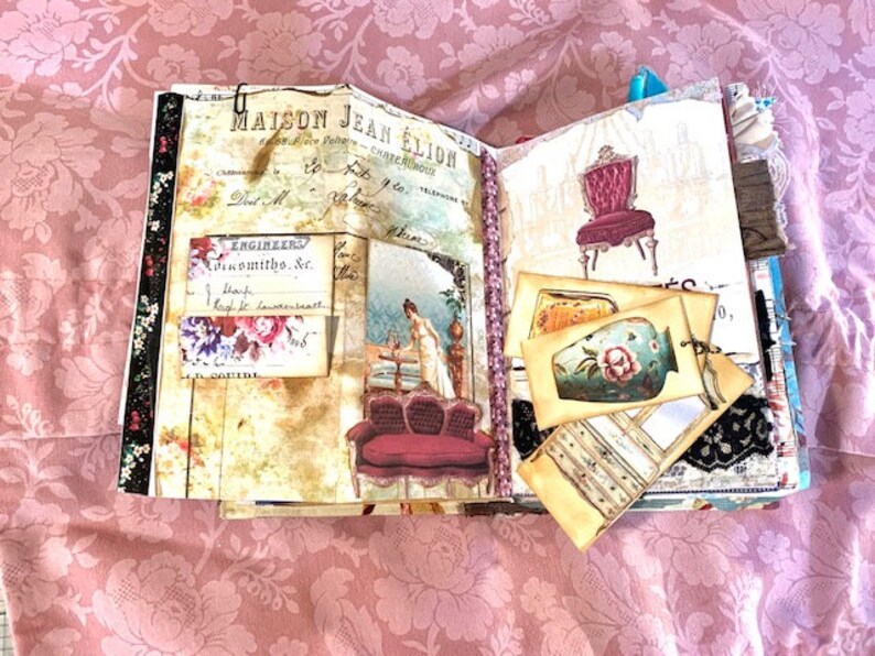 May include: Open journal with vintage-style pages. The pages feature illustrations of furniture, floral accents, and handwritten text. The journal is adorned with decorative elements, including lace and ribbon, and a metal clip.