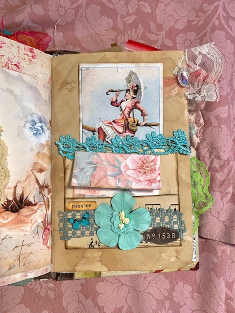 May include: A handmade journal with a vintage aesthetic. Features a collage with a Victorian illustration, teal lace, a paper flower, and the word "passion". The journal has a floral patterned background.