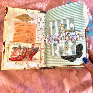 May include: Open journal with vintage-style pages. One page features a colorful illustration of a dog in a red shoe with a rose. The other page has a striped pattern with a decorative card and butterfly embellishments.