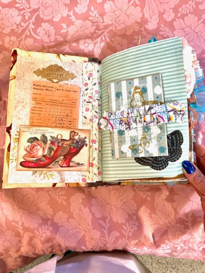 May include: Open journal with vintage-style pages. One page features a colorful illustration of a dog in a red shoe with a rose. The other page has a striped pattern with a decorative card and butterfly embellishments.