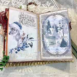 May include: Open scrapbook with a Christmas theme. The left page features a deer wearing a Santa hat and the words "Merry Christmas." The right page shows a winter scene with a house, trees, and a gate. The pages are trimmed with gold fringe.