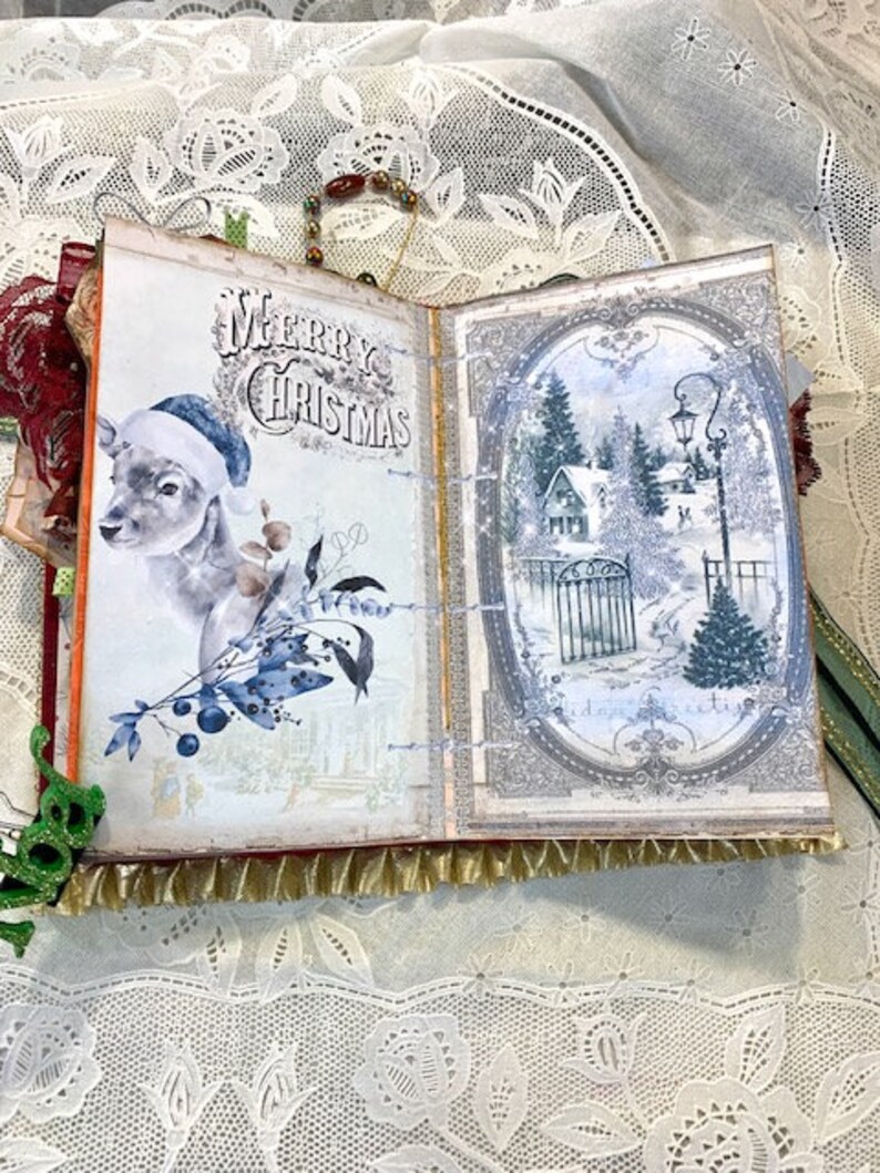 May include: Open scrapbook with a Christmas theme. The left page features a deer wearing a Santa hat and the words "Merry Christmas." The right page shows a winter scene with a house, trees, and a gate. The pages are trimmed with gold fringe.