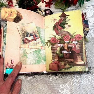 May include: Open journal with Christmas-themed illustrations. One page features a winter scene with a red barn, while the other depicts a decorated fireplace with stockings. The journal is adorned with festive embellishments and a Santa Claus image.