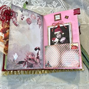 May include: Open journal with Christmas-themed pages. The left page features watercolor illustrations of ornaments and a poinsettia. The right page has a photo of a child in a Santa hat and a striped pocket. Gold fringe trims the bottom.