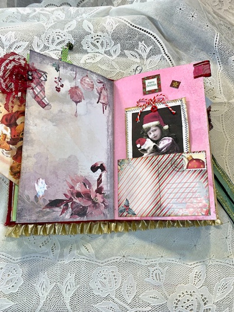 May include: Open journal with Christmas-themed pages. The left page features watercolor illustrations of ornaments and a poinsettia. The right page has a photo of a child in a Santa hat and a striped pocket. Gold fringe trims the bottom.