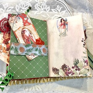 May include: Open journal with Christmas-themed pages. The left page is green with a white diamond pattern and a pocket with a pinecone design. The right page has a floral and angel illustration. A Santa tag is attached.