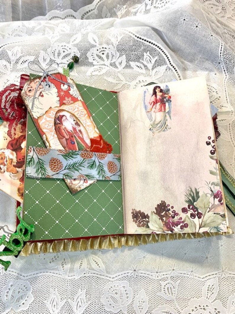 May include: Open journal with Christmas-themed pages. The left page is green with a white diamond pattern and a pocket with a pinecone design. The right page has a floral and angel illustration. A Santa tag is attached.