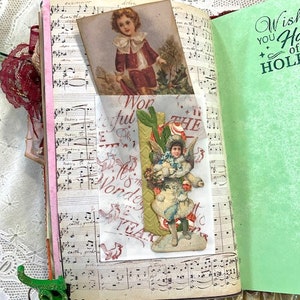 May include: Open journal with vintage pages featuring sheet music and decorative inserts. A small card shows a child in a red coat. A green page reads "Wish You Happy Holidays". A green binder clip is attached.