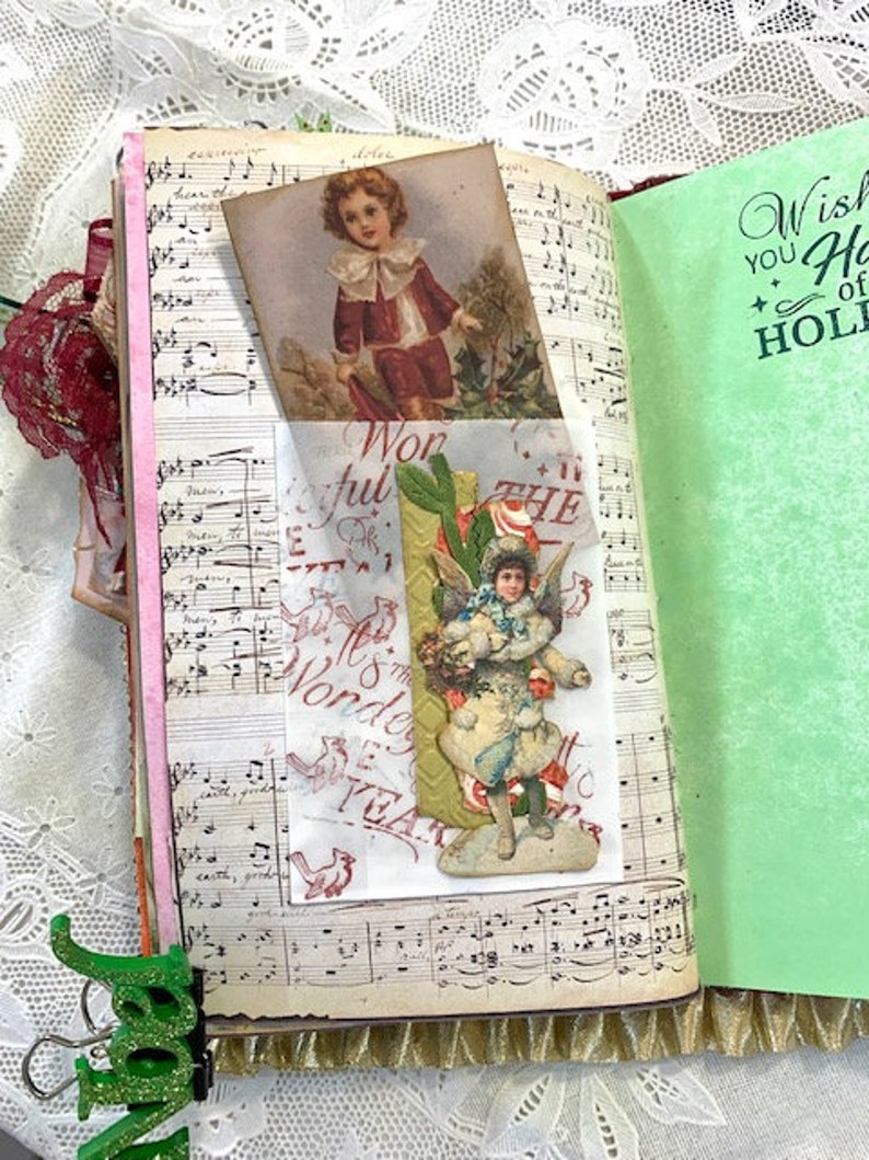 May include: Open journal with vintage pages featuring sheet music and decorative inserts. A small card shows a child in a red coat. A green page reads "Wish You Happy Holidays". A green binder clip is attached.
