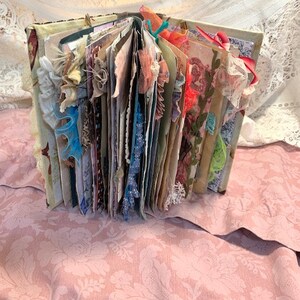 May include: An open, handmade journal with decorative pages and embellishments. The journal's pages are visible, featuring various colors, textures, and decorative elements like lace, ribbons, and fabric scraps. The cover is visible, with a floral pattern. The journal is resting on a pink floral fabric.