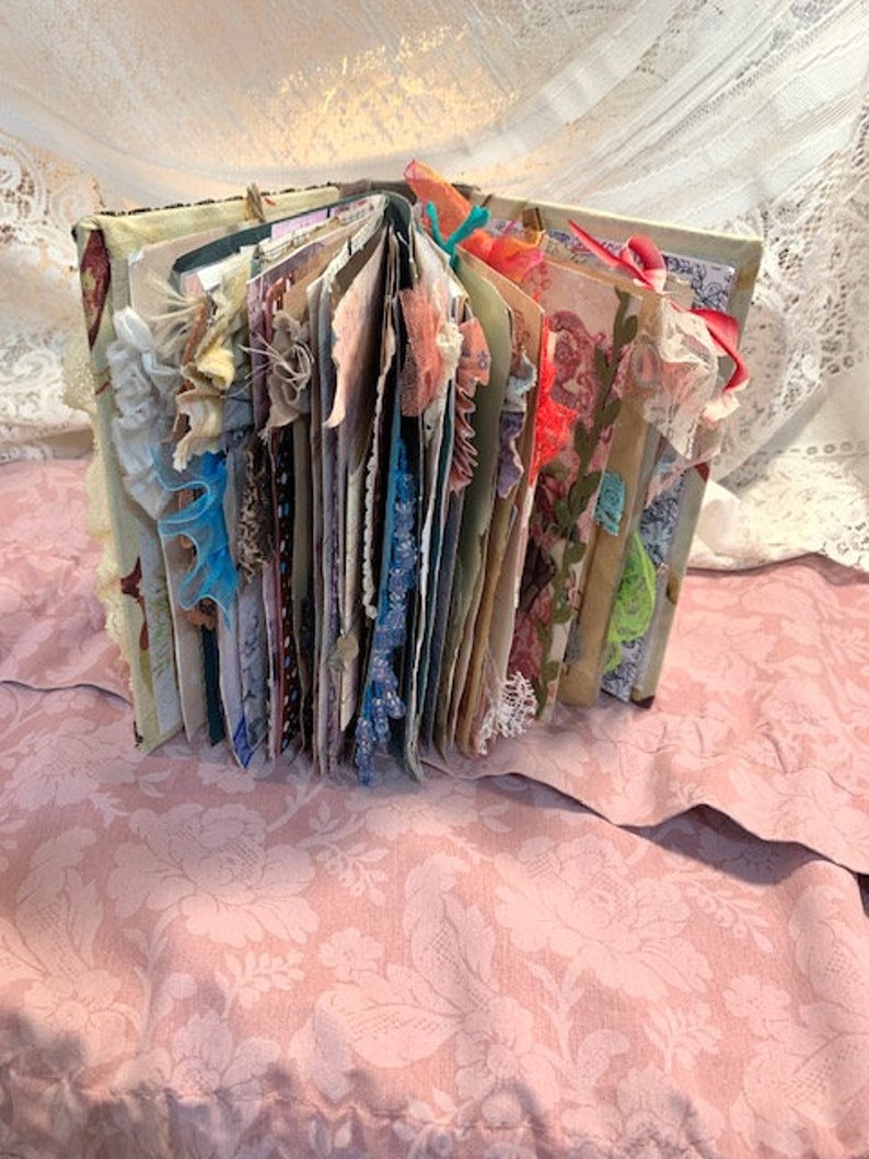 May include: An open, handmade journal with decorative pages and embellishments. The journal's pages are visible, featuring various colors, textures, and decorative elements like lace, ribbons, and fabric scraps. The cover is visible, with a floral pattern. The journal is resting on a pink floral fabric.