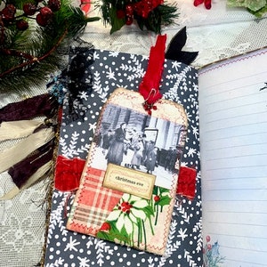 May include: A handmade journal with a gray snowflake pattern cover. A tag with a vintage photo and the words "Christmas Eve" is attached with a red ribbon. The journal is open to lined pages.