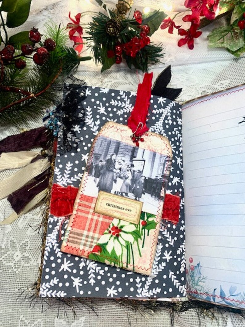 May include: A handmade journal with a gray snowflake pattern cover. A tag with a vintage photo and the words "Christmas Eve" is attached with a red ribbon. The journal is open to lined pages.