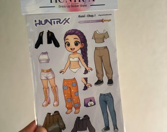 Könnte beinhalten: Ein Anzieh-Stickerbogen mit einer Cartoonfigur mit lila Haaren und verschiedenen Kleidungsoptionen. Der Bogen enthält Jacken, Oberteile, Hosen, Shorts, Stiefel und Accessoires. Der Text "KPOP DEMON HUNTERS HUNTR/X Dress Up Sticker Sheet" ist sichtbar.