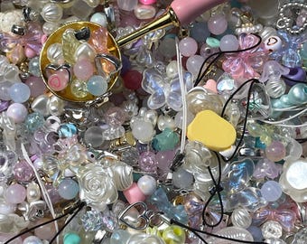 May include: A large assortment of colorful beads, charms, and other decorative elements, including hearts, flowers, butterflies, and seashells. The beads are arranged in a random pattern and are mostly pastel colors, such as pink, blue, and purple. There is a pink and gold scoop in the image.
