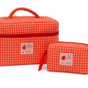 May include: Two-piece set of red and white gingham patterned cosmetic bags. The larger bag has a handle and a red zipper. The smaller bag has a wooden ring attached. Both bags have a label with a ladybug illustration and text.