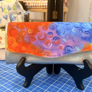 May include: Silver clutch wallet with a flap featuring a colorful swirl pattern in orange, purple, and blue. The wallet is displayed on a black stand against a blue grid background.