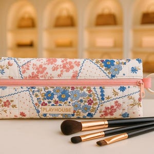 May include: A floral print makeup bag with a pink zipper and pull, and a matching handle. The bag is next to three makeup brushes with black handles and gold-colored metal. The bag has the word "PLAYHOUSE" on it.