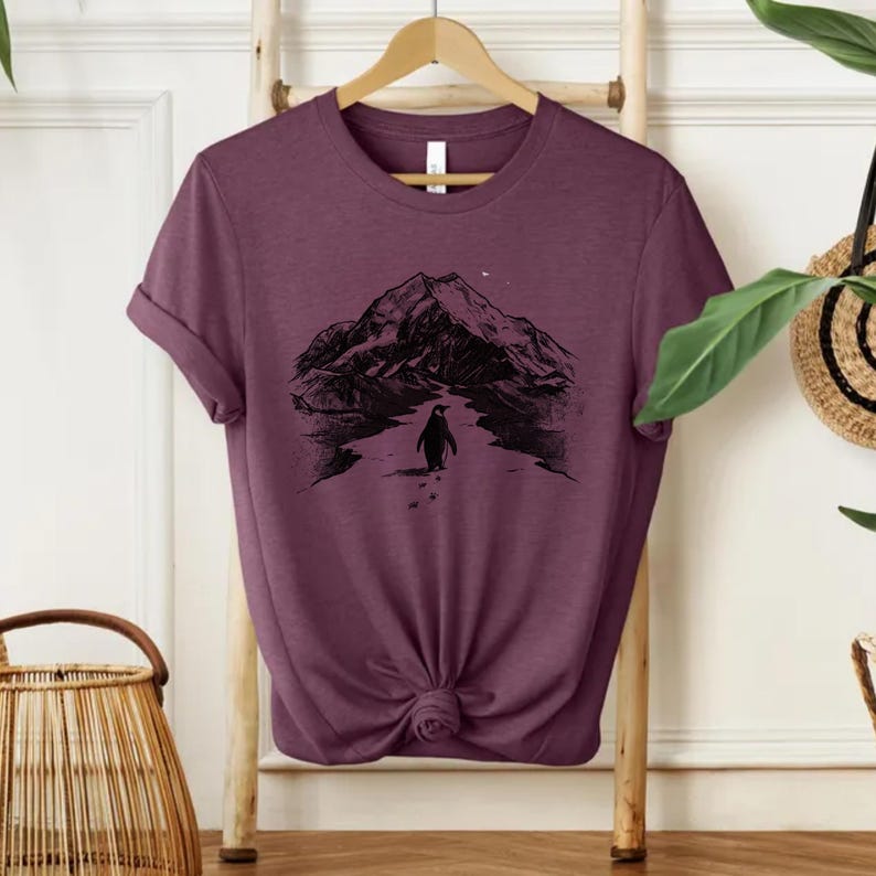 May include: A maroon t-shirt featuring a black graphic of a penguin walking towards a mountain range under a starry sky. The shirt has short sleeves and a knotted front. The design is centered on the front of the shirt.