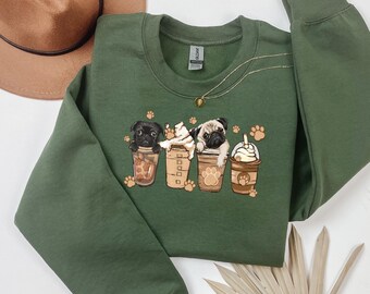 Pode incluir: Um moletom verde-oliva com um gráfico de quatro pugs em xícaras de café. Os pugs são pretos e castanhos, com chantilly e pegadas. Um colar de ouro e um chapéu marrom também são visíveis.