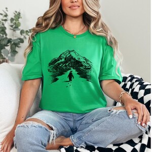 May include: A green t-shirt featuring a black graphic of a penguin walking towards a mountain range. The penguin is walking on a path with footprints. The shirt is a solid green color.