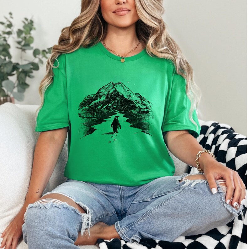 May include: A green t-shirt featuring a black graphic of a penguin walking towards a mountain range. The penguin is walking on a path with footprints. The shirt is a solid green color.