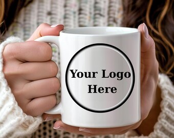 May include: White ceramic mug with a handle, featuring the text "Your Logo Here" inside a black circle. The mug is held by a person wearing a white sweater.