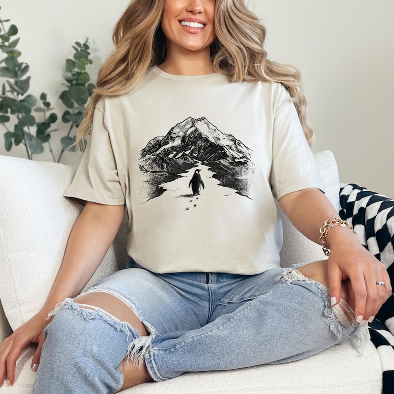 May include: A beige t-shirt featuring a black and white graphic of a penguin walking towards a mountain. The penguin is depicted in the center of the shirt, with a mountain range in the background. The shirt is made of a soft material.