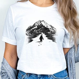 May include: White t-shirt featuring a black and white illustration of a penguin walking towards a mountain range. The graphic shows the penguin's footprints in the snow. The shirt is made of a soft material.