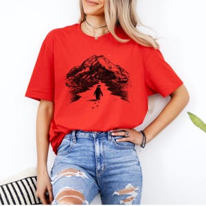 May include: A red t-shirt featuring a black graphic of a penguin walking towards a mountain range. The penguin is leaving footprints in the snow. The shirt is paired with blue jeans.