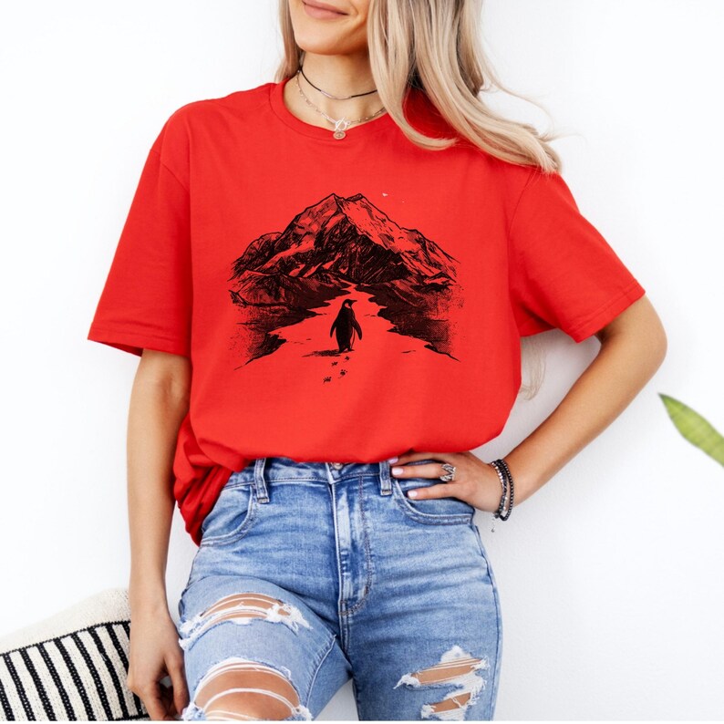 May include: A red t-shirt featuring a black graphic of a penguin walking towards a mountain range. The penguin is leaving footprints in the snow. The shirt is paired with blue jeans.