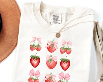 May include: Cream-colored t-shirt featuring a strawberry design. The strawberries are red with green leaves and pink bows. The shirt has a tag that says "COMFORT COLORS". A gold necklace is draped across the shirt.