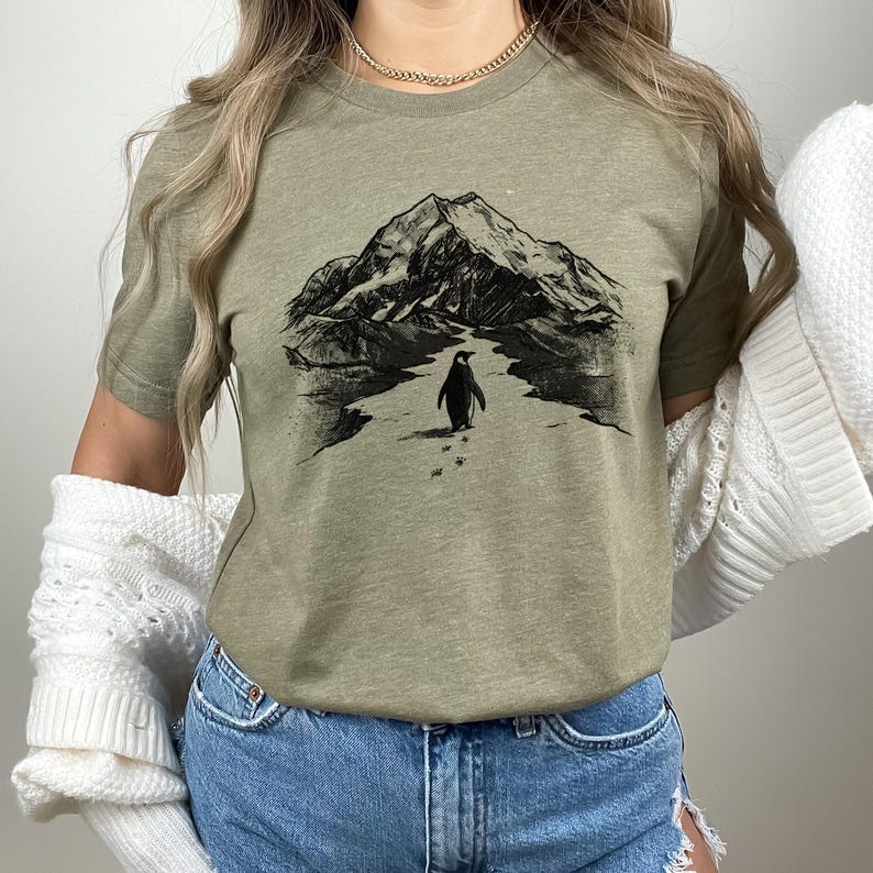 May include: Olive green t-shirt featuring a black and white illustration of a penguin walking towards a mountain. The graphic includes the penguin's footprints and a detailed mountain range in the background. The shirt has short sleeves and a crew neck.