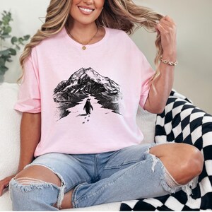 May include: A light pink t-shirt featuring a black and white illustration of a mountain range and a penguin walking on snow. The shirt is a crew neck style.