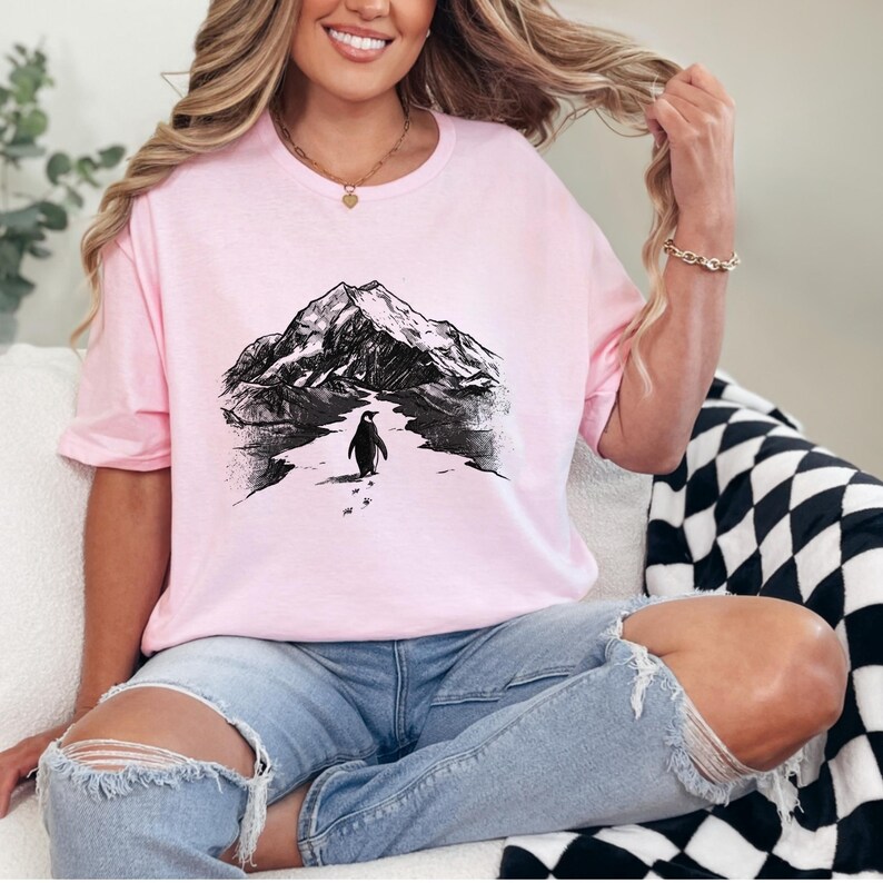 May include: A light pink t-shirt featuring a black and white illustration of a mountain range and a penguin walking on snow. The shirt is a crew neck style.