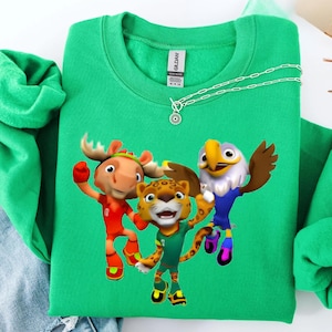 May include: A green sweatshirt featuring a graphic of three cartoon characters: a moose, a leopard, and an eagle. The moose wears red, the leopard wears green, and the eagle wears blue. A silver necklace with a pendant is draped across the sweatshirt.