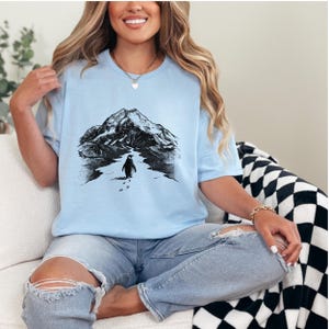 May include: Light blue t-shirt featuring a black and white graphic of a penguin walking towards a mountain range. The design includes the penguin's footprints in the snow. The shirt is made of a soft material.