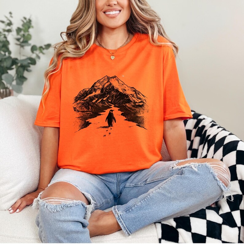 May include: Bright orange t-shirt featuring a black and white graphic of a penguin walking towards a mountain. The graphic is centered on the shirt. The person is wearing light blue jeans with ripped knees.