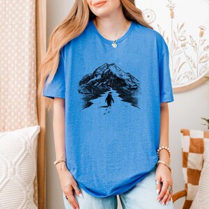 May include: A blue t-shirt featuring a black and white graphic of a penguin walking towards a mountain range. The penguin is depicted with footprints in the snow. The shirt has a crew neck and short sleeves.