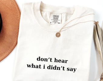 May include: Cream-colored t-shirt with the phrase "don't hear what i didn't say" printed in black. The shirt has a crew neck and short sleeves. A gold necklace with a pendant rests on the shirt. The shirt is folded on a white surface.