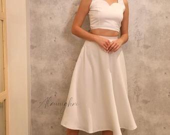Puede incluir: Un conjunto blanco de dos piezas con un top corto con escote corazón y una falda midi a juego. El conjunto se combina con tacones negros. El fondo es una pared gris texturizada.