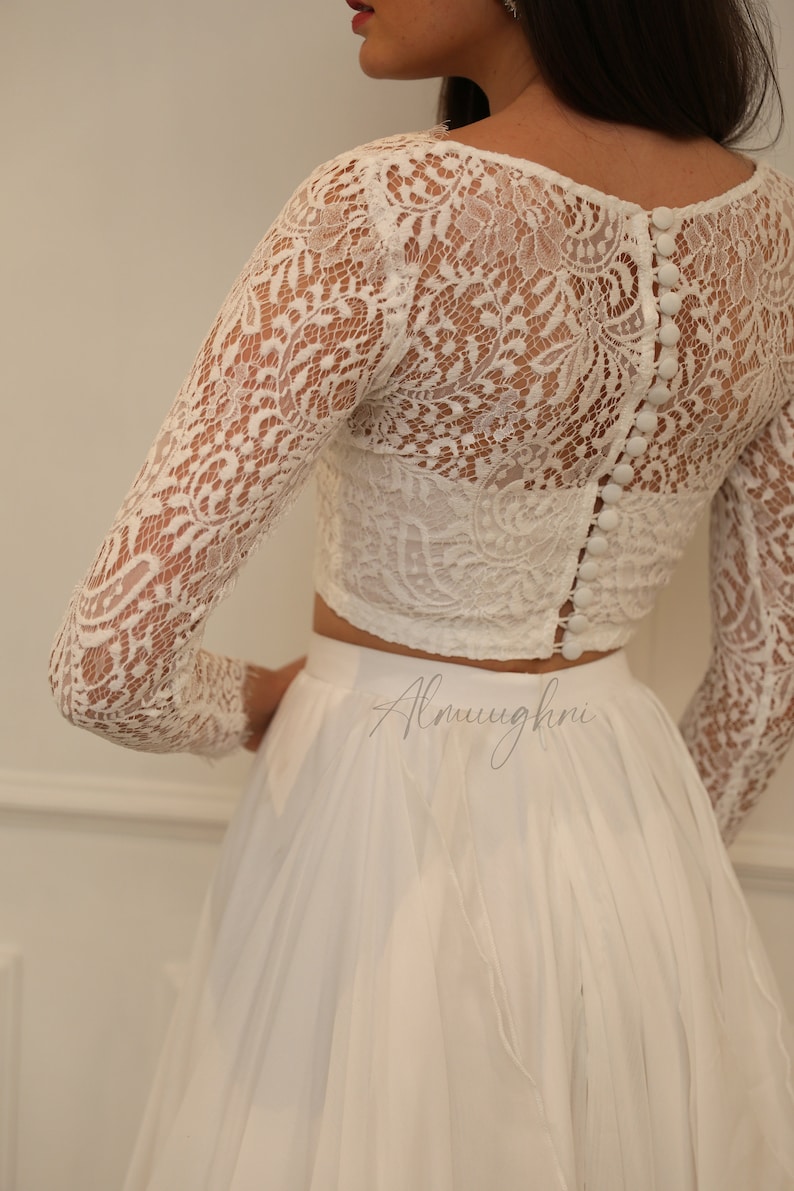 May include: A white lace crop top with long sleeves and a button closure down the back. The top is paired with a white tulle skirt. The skirt is flowing and has a ruffled hem. The text 'Almughni' is visible on the skirt.