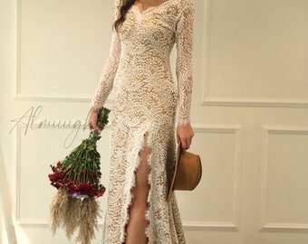 May include: A white lace wedding dress with long sleeves and a thigh-high slit. The dress has a V-neckline and a fitted silhouette. The model is holding a bouquet of red flowers and a brown hat.