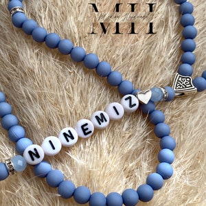 Op de afbeelding: Close-up van twee lichtblauwe kralenarmbanden. E&eacute;n armband spelt de naam "NINEMIZ" in witte letterkralen. De armbanden hebben zilveren accenten en een kleine hartbedel. De achtergrond is een lichtbruin, getextureerd oppervlak.