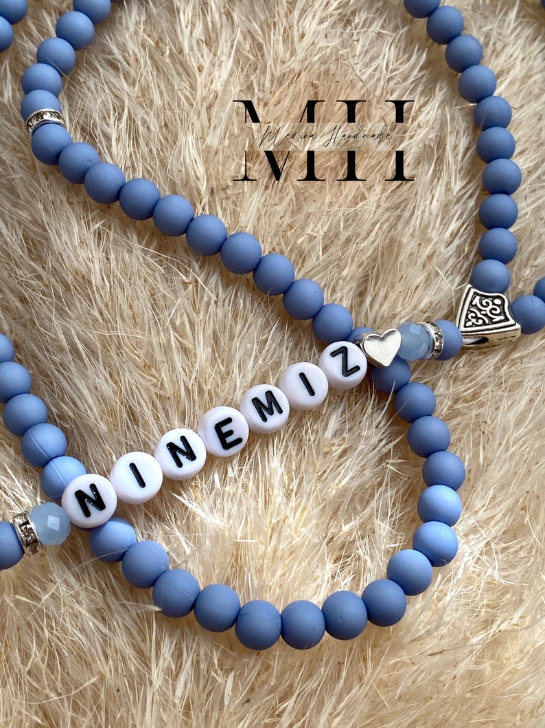 Op de afbeelding: Close-up van twee lichtblauwe kralenarmbanden. E&eacute;n armband spelt de naam "NINEMIZ" in witte letterkralen. De armbanden hebben zilveren accenten en een kleine hartbedel. De achtergrond is een lichtbruin, getextureerd oppervlak.