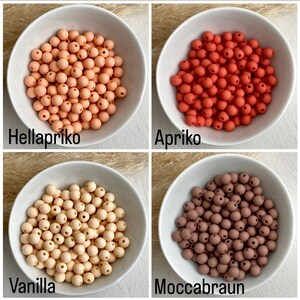 Op de afbeelding: Vier kommen met ronde kralen in verschillende kleuren: licht perzik, roodachtig oranje, cr&egrave;me en bruin. De kommen zijn gelabeld met "Hellapriko", "Apriko", "Vanilla" en "Moccabraun".
