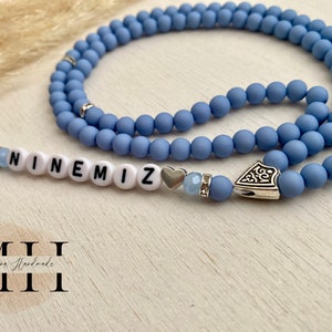 Op de afbeelding: Een lichtblauwe kralenketting met het woord "NINEMIZ" gespeld in witte letterkralen. De ketting heeft zilverkleurige accenten, waaronder een hartvormige bedel en een decoratieve hanger.